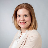 Portrait of a smiling woman with shoulder-length light brown hair, wearing a beige blazer and a white blouse against a neutral background.